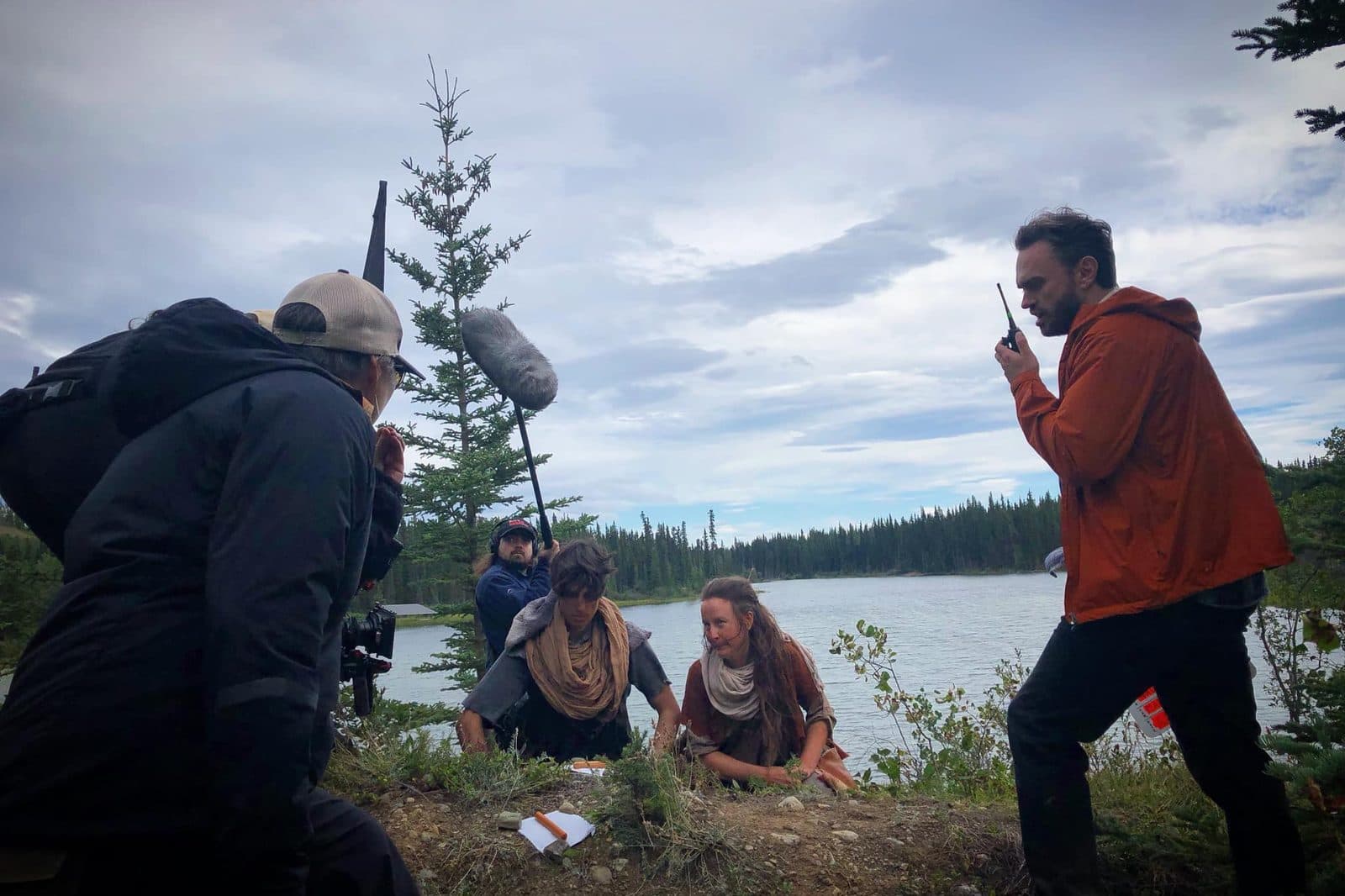 Kryshan Randel on a walkie-talkie by a lake while two actors kneel for a take, with boom and camera beside them.